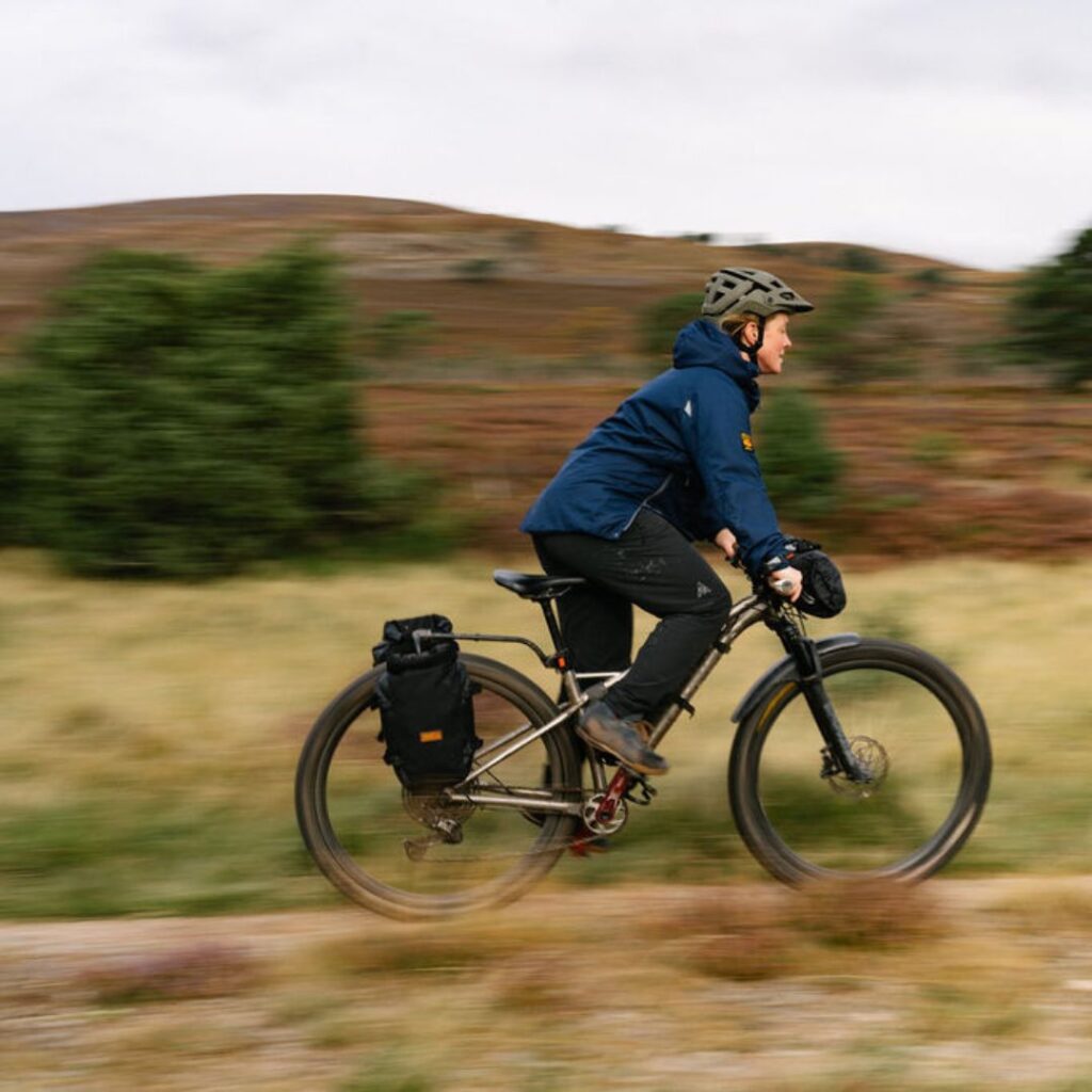 Woman in sustainable gear riding a bike along a stony path surrounded by grassland 