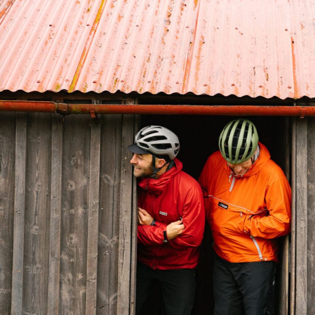 Two men in bright red and orange sustainable gear quarter zips and bike helmets peering out of a cabin door